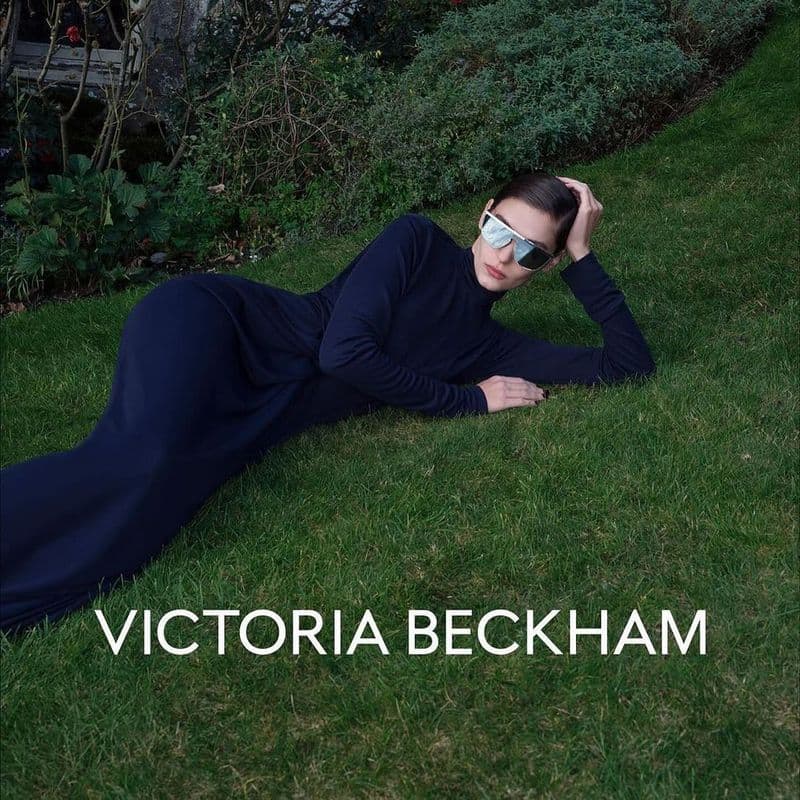 Model in dark sunglasses and long dark dress reclining on a green lawn for Victoria Beckham