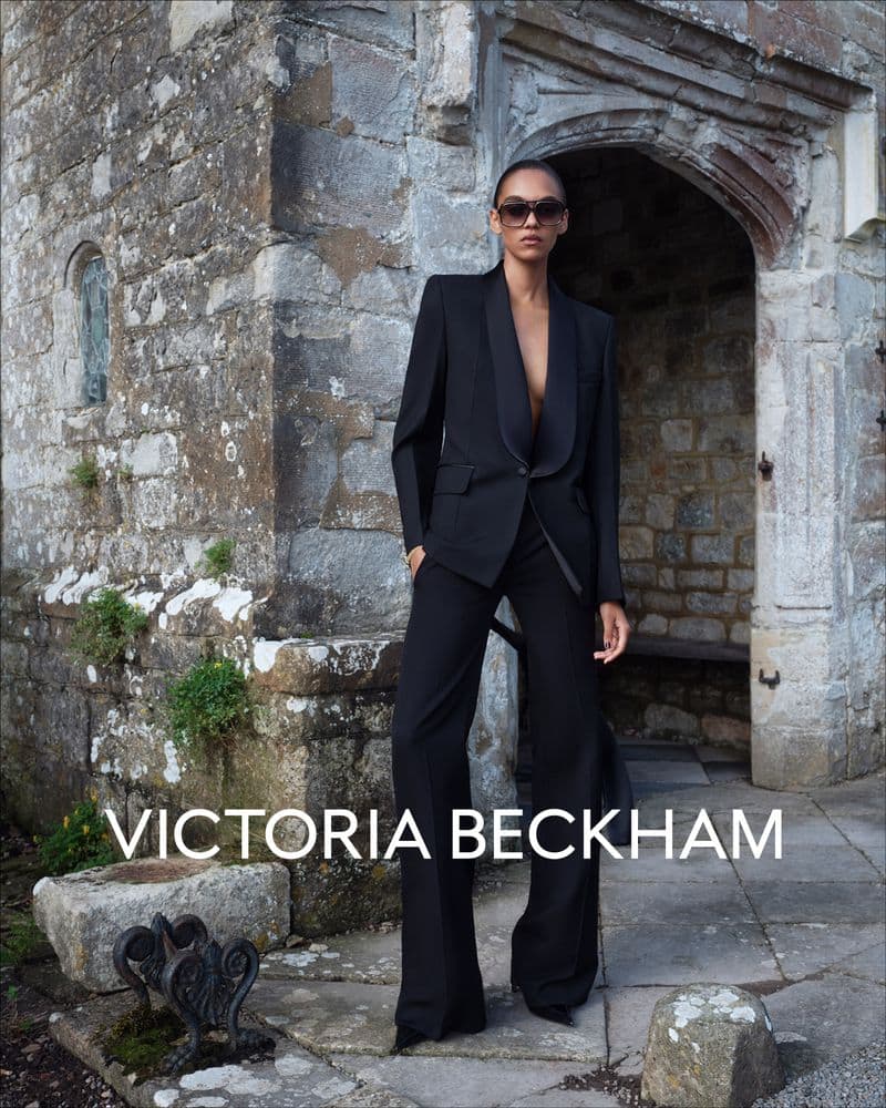 Model wearing a black pantsuit and sunglasses standing in front of an ancient stone archway for Victoria Beckham.