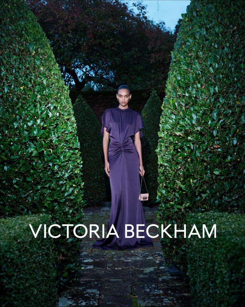 Model in long purple dress holding small handbag framed by manicured topiary hedges at twilight