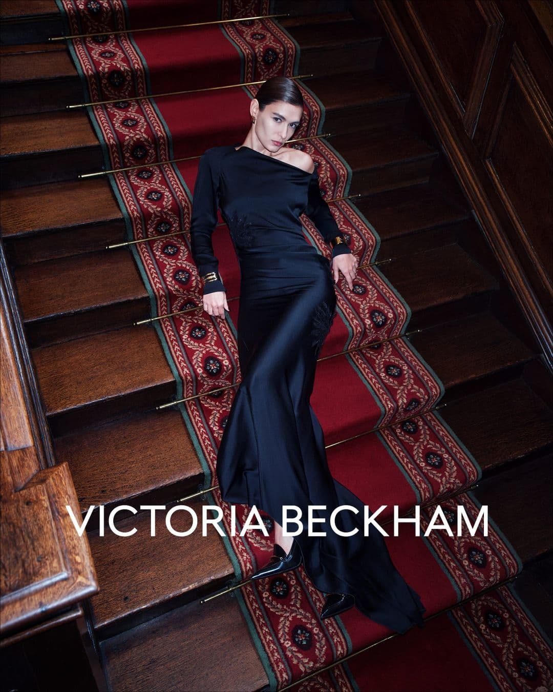 Model in a black satin gown lying on a wooden staircase with a patterned red carpet runner for Victoria Beckham