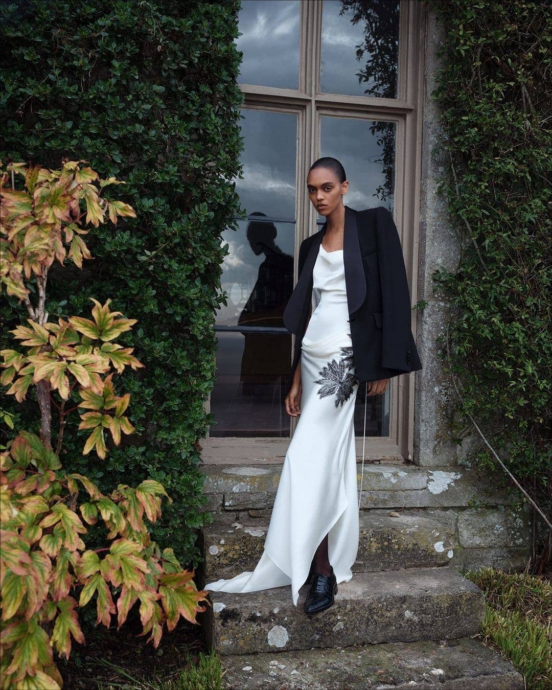 Model standing on stone steps in a white slip dress with black blazer outdoors near foliage
