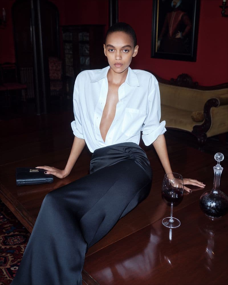 Model seated on a dark wooden table wearing a white button-down shirt and black skirt in an interior room.