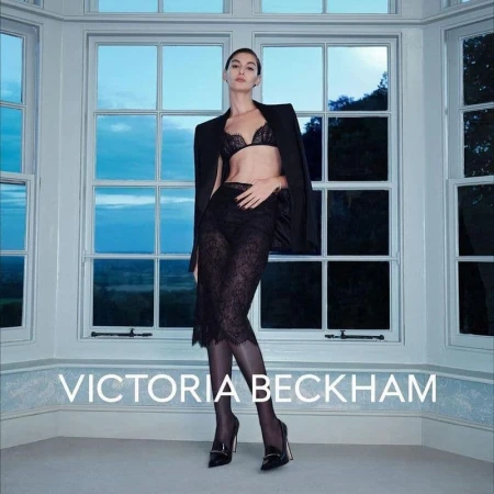 Model wearing a black lace bra, skirt, blazer, and dark tights standing in front of large multi-pane windows.