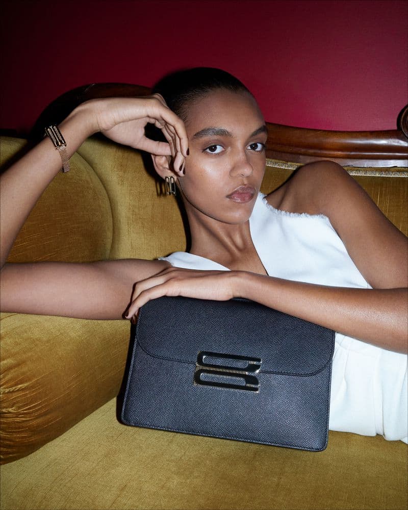 Model reclining on a yellow tufted sofa holding a black handbag for Victoria Beckham
