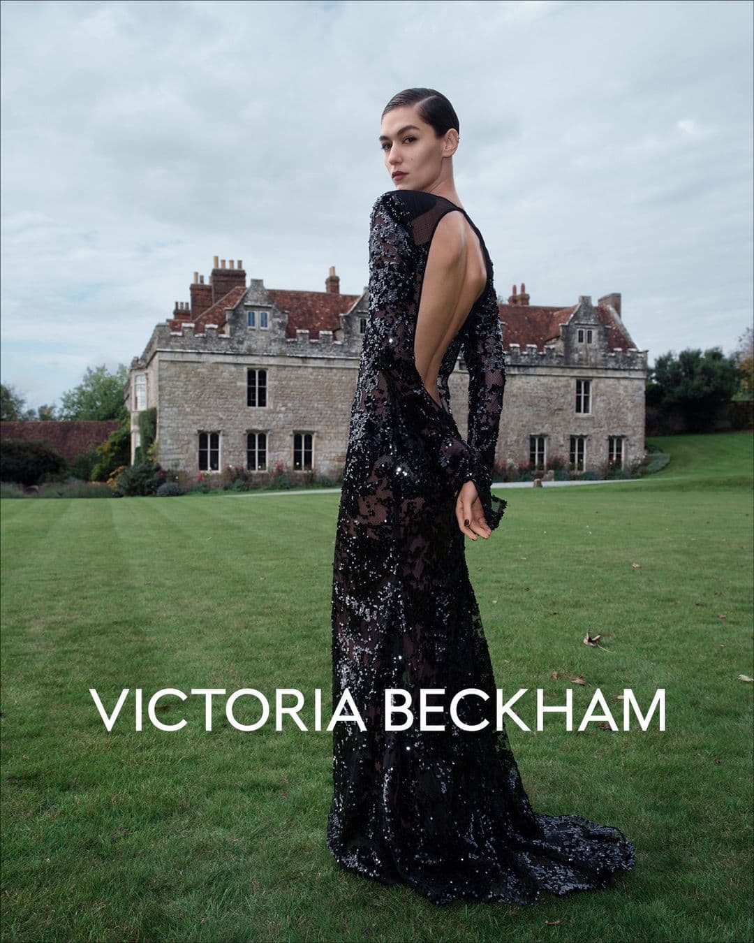 Model in a black sequin gown looking back on a lawn before a historic English country house under an overcast sky.