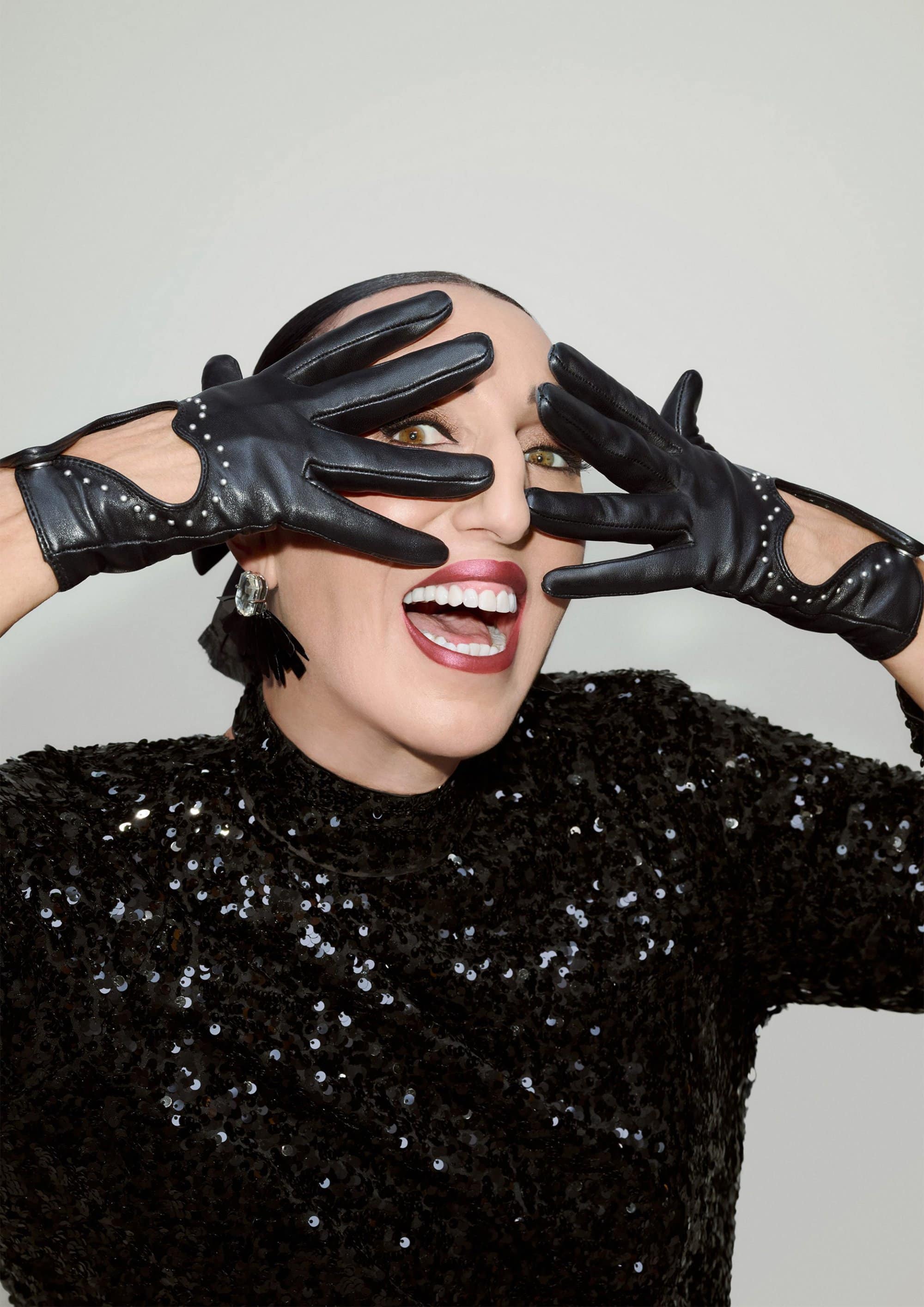 Model Rossy de Palma wearing black leather gloves over her face and a black sequined top against a white studio background.
