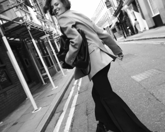 Woman walking down a city street wearing a tailored blazer and carrying a dark handbag, captured in motion.