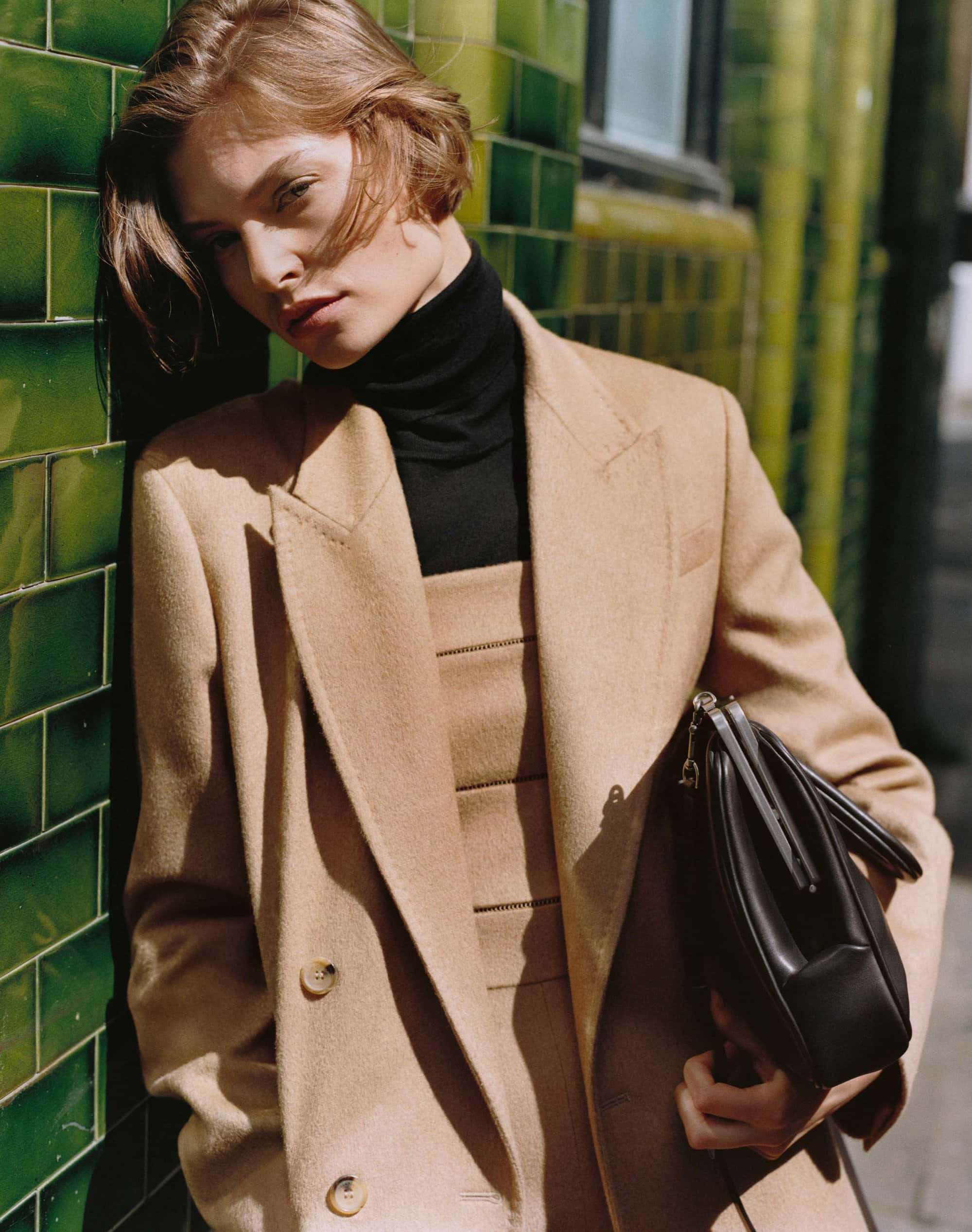 Model leaning against green tiled wall wearing a camel coat and holding a black handbag for Max Mara