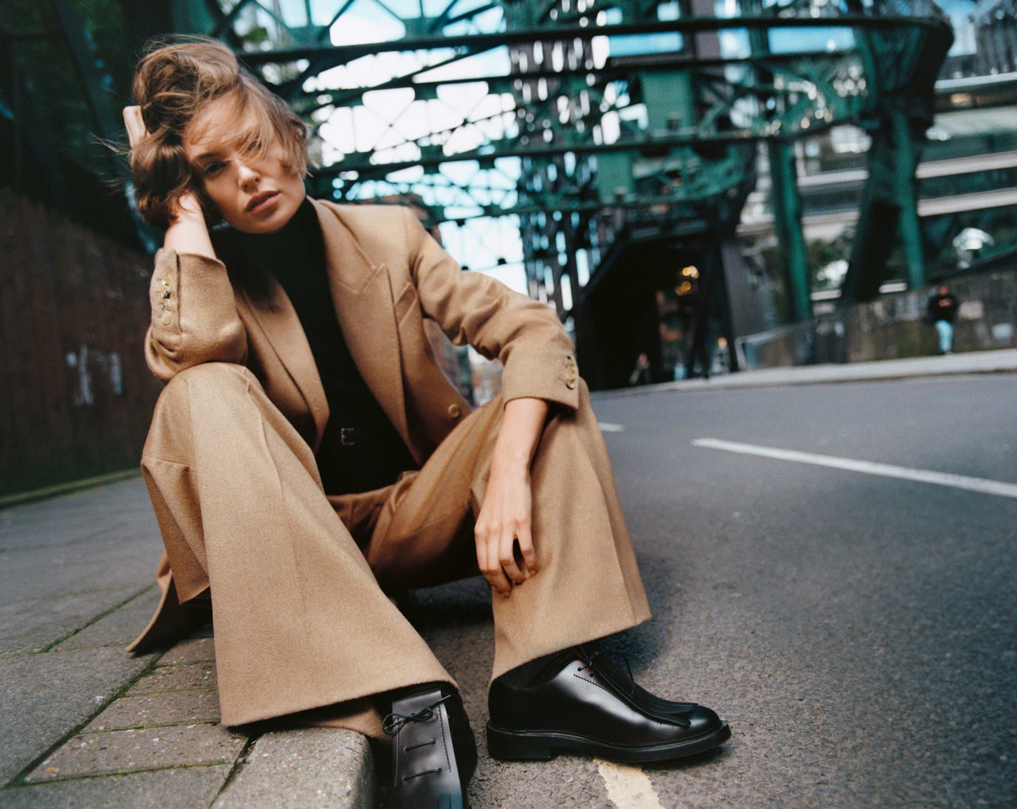 Model Bibi Breslin wearing a camel suit and black shoes sitting on an urban street for Max Mara Fall 2024 campaign