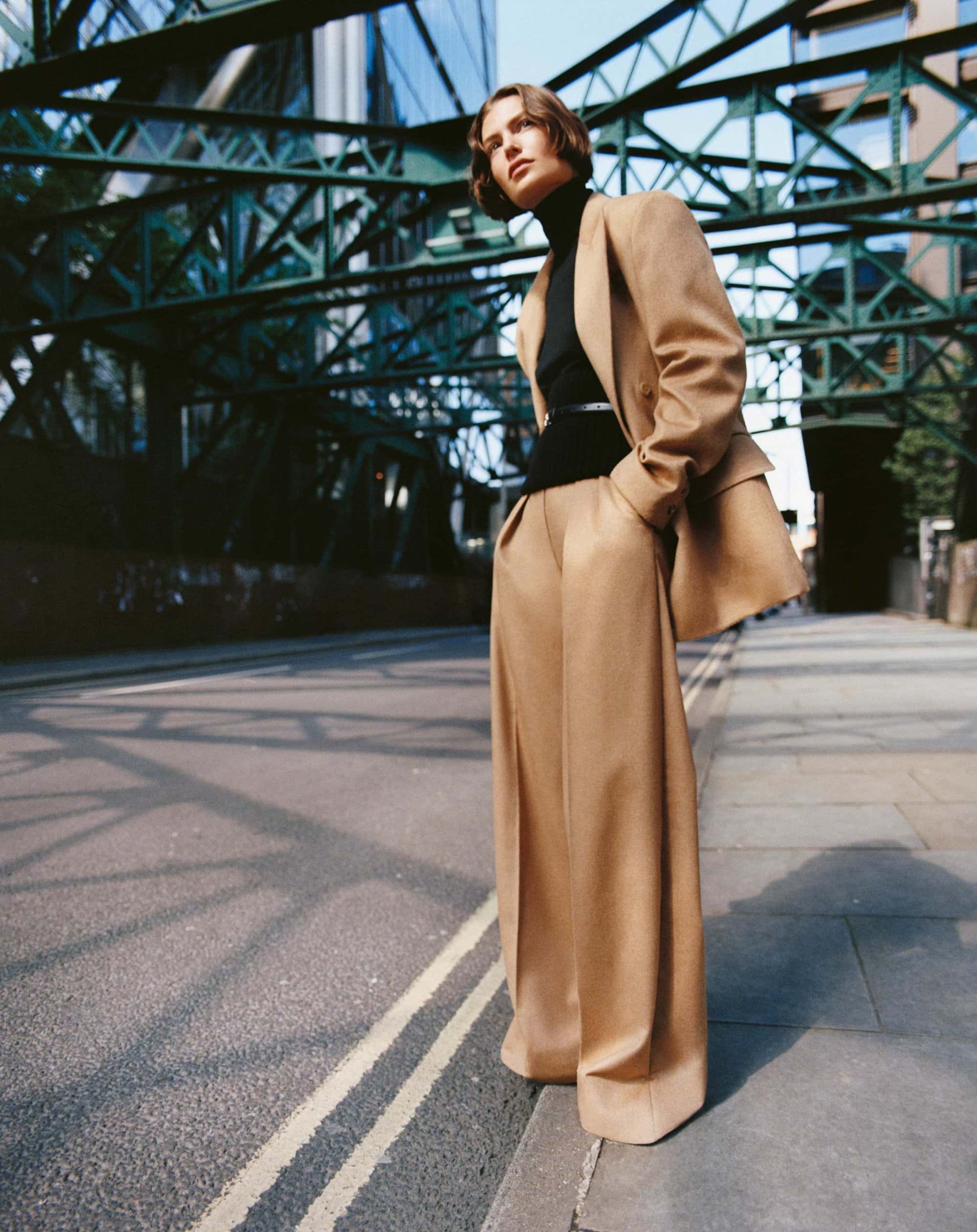 Model standing on a city street under an iron bridge wearing a camel suit and black turtleneck