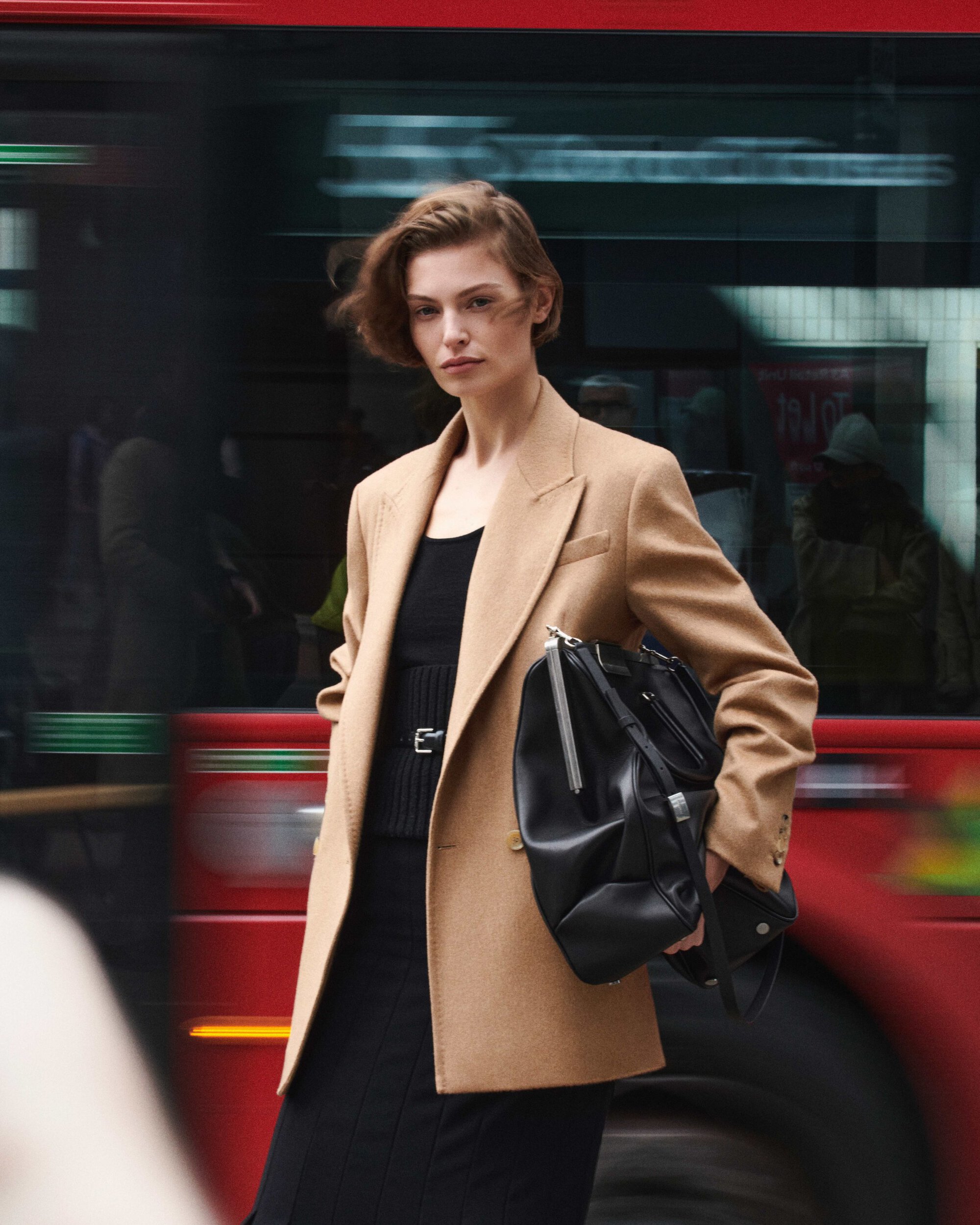 Model wearing a camel coat and black top holding a black leather handbag in front of a passing red bus