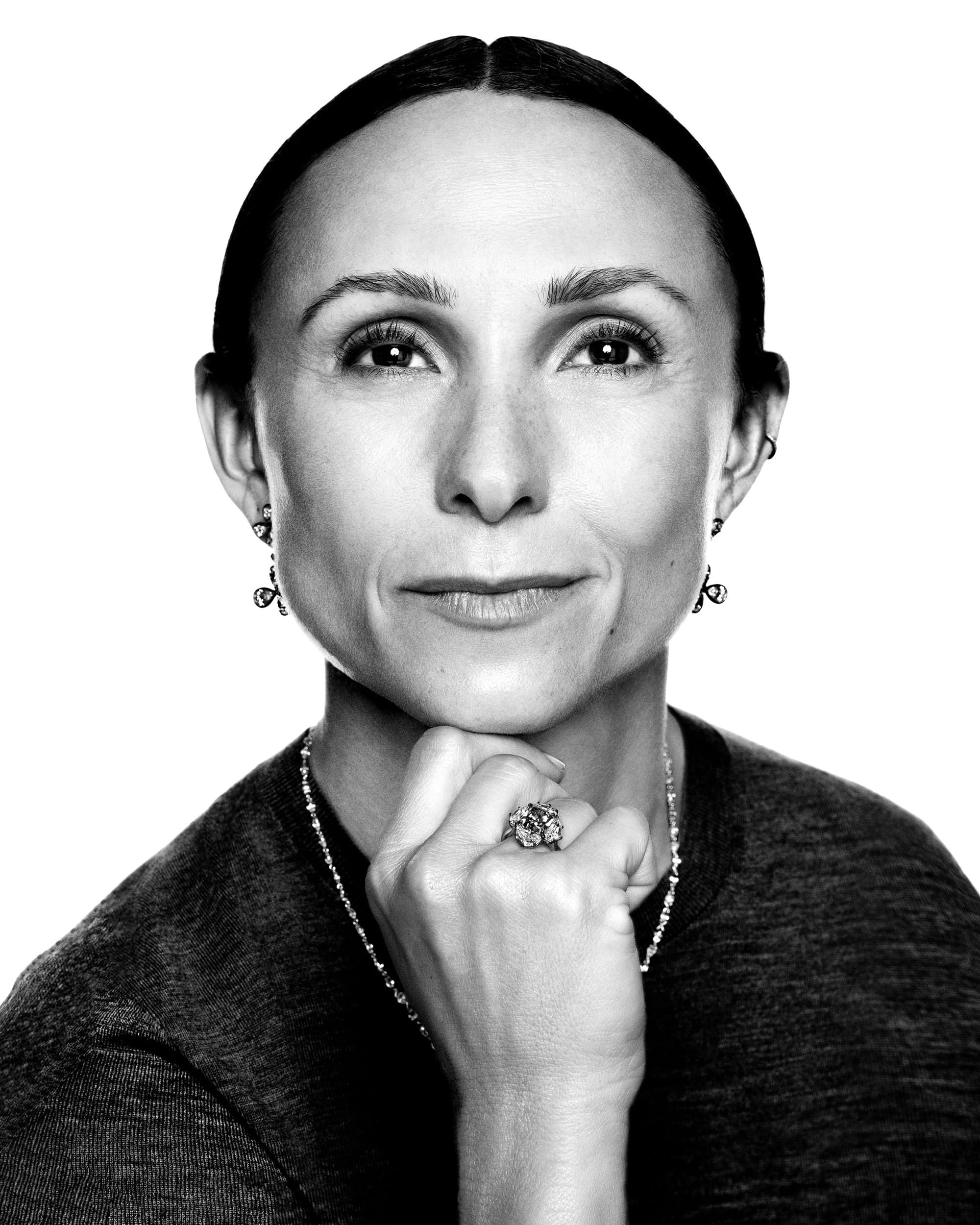 Black and white close-up portrait of a woman resting her chin on her hand, wearing delicate jewelry against a plain background.
