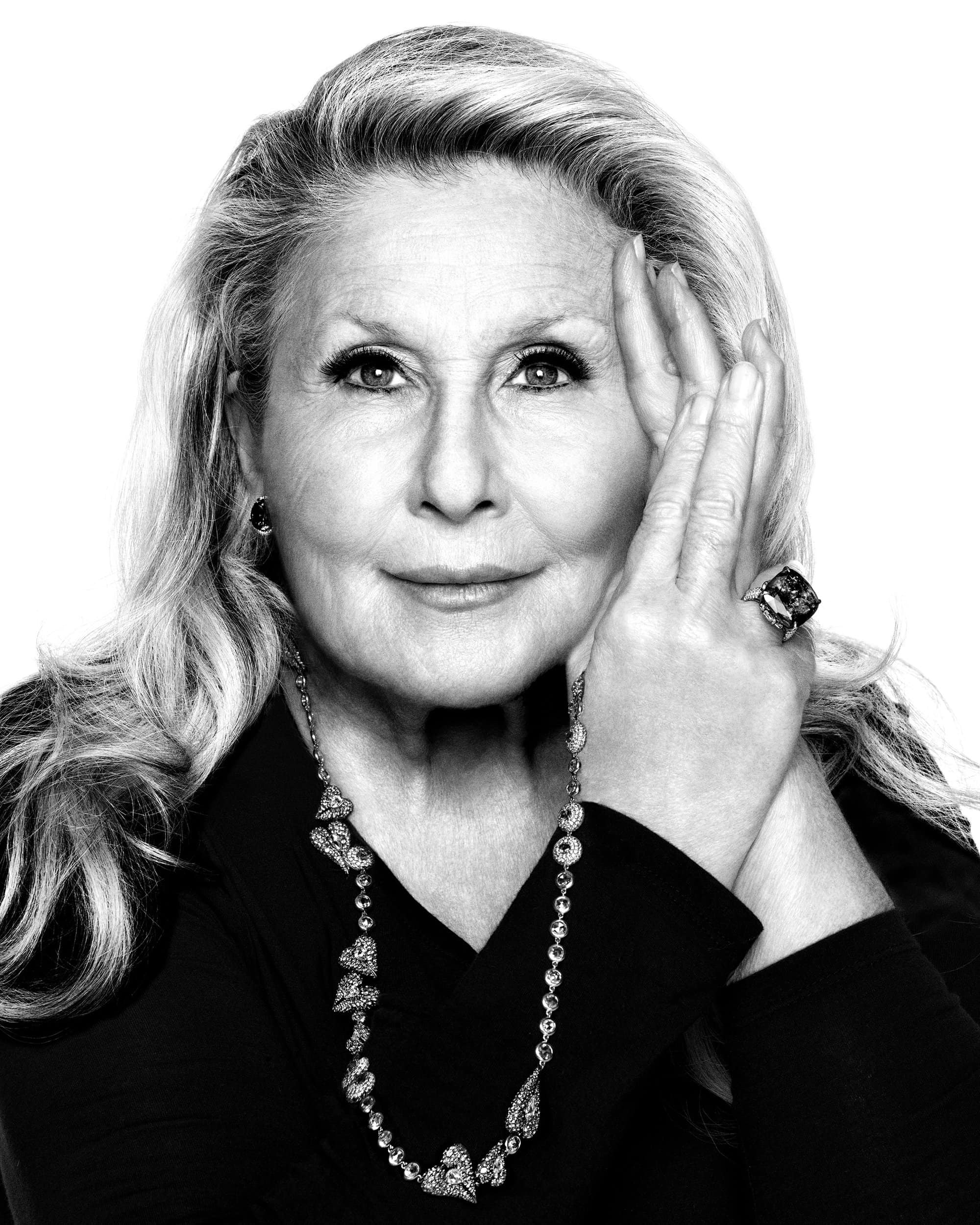 Close-up monochrome portrait of an older woman wearing diamond jewelry against a plain white seamless background.