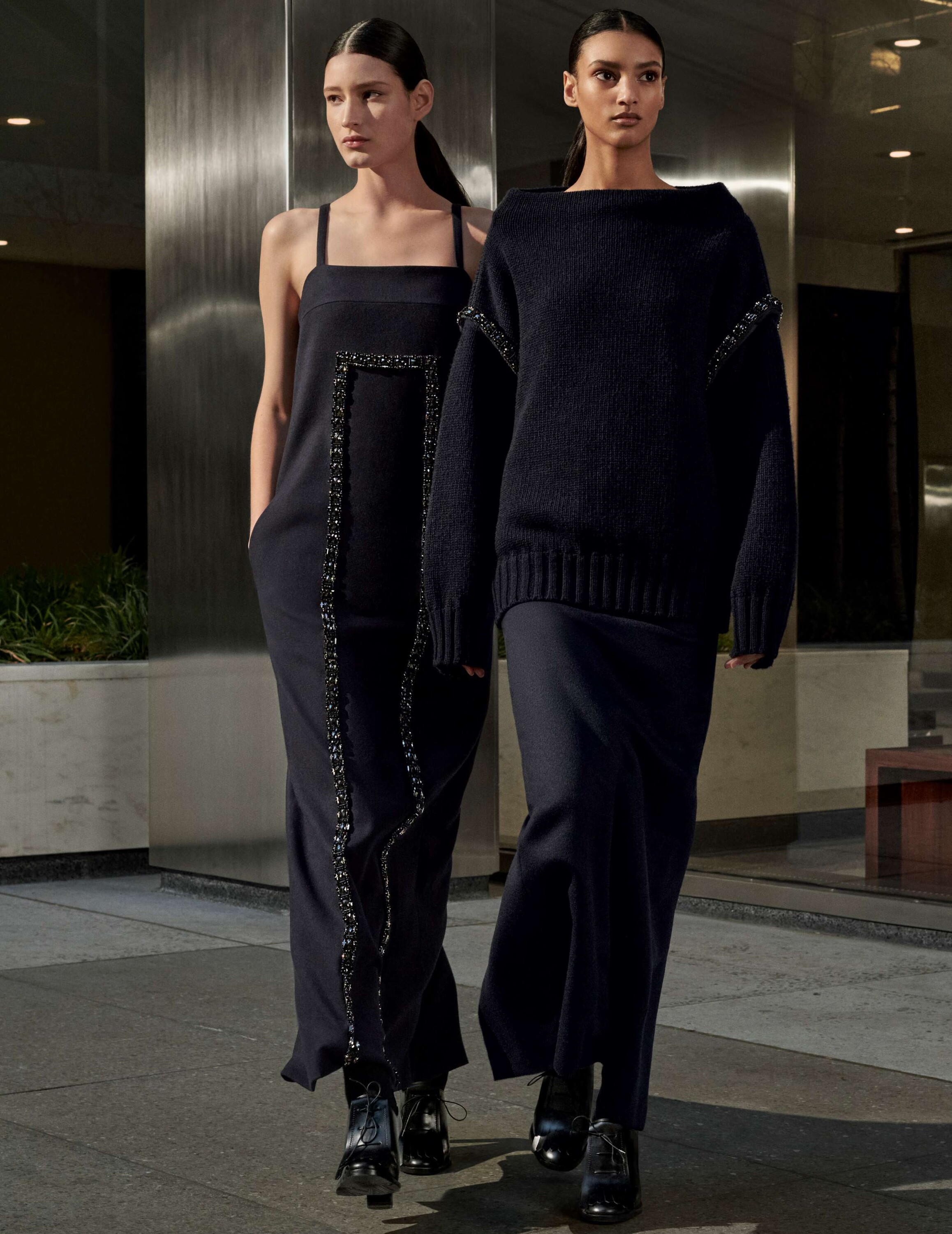 Two female models in dark designer outfits walking together outdoors near a modern metallic building exterior.