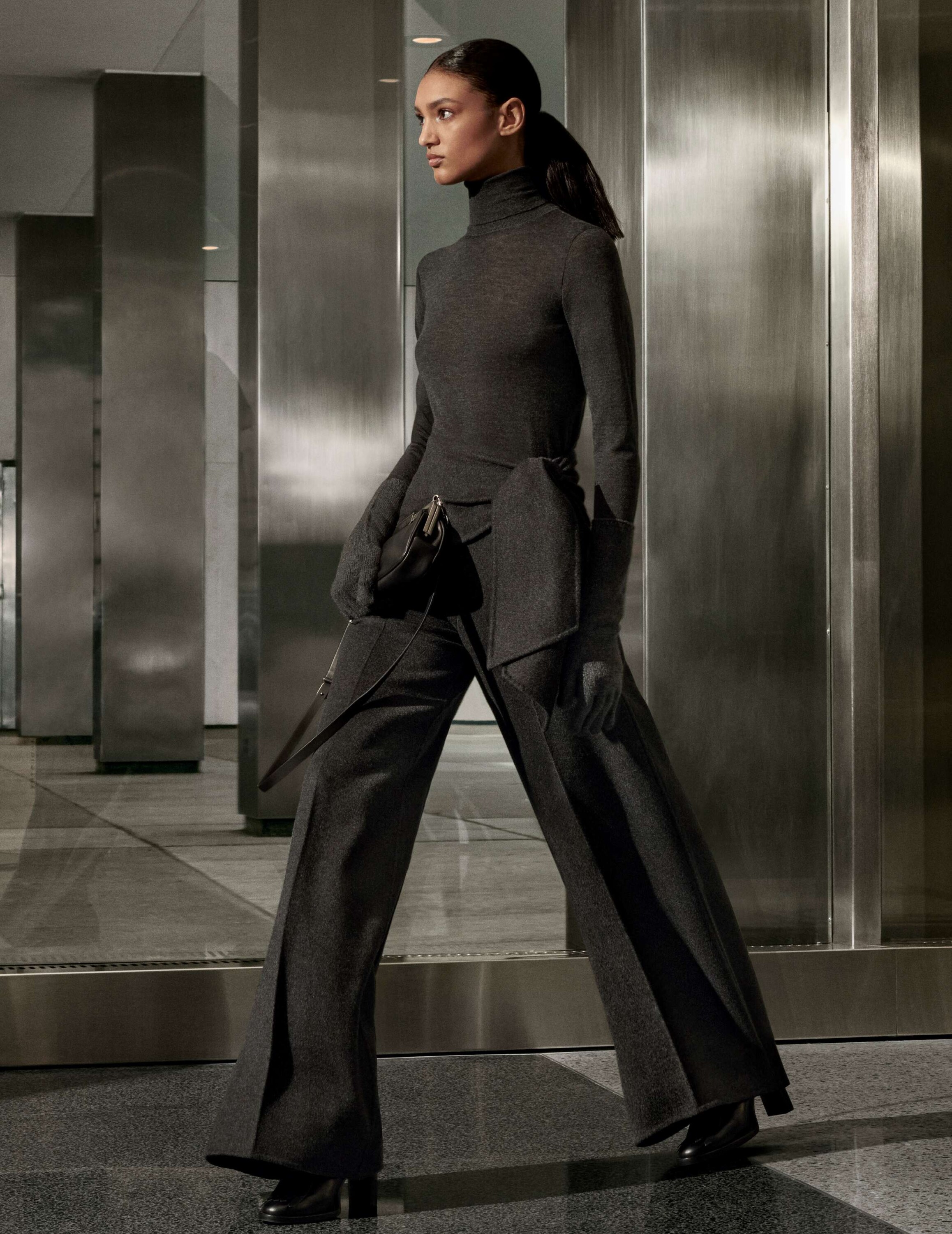 Model in charcoal grey turtleneck and wide trousers holding a handbag against a modern metallic architectural interior.