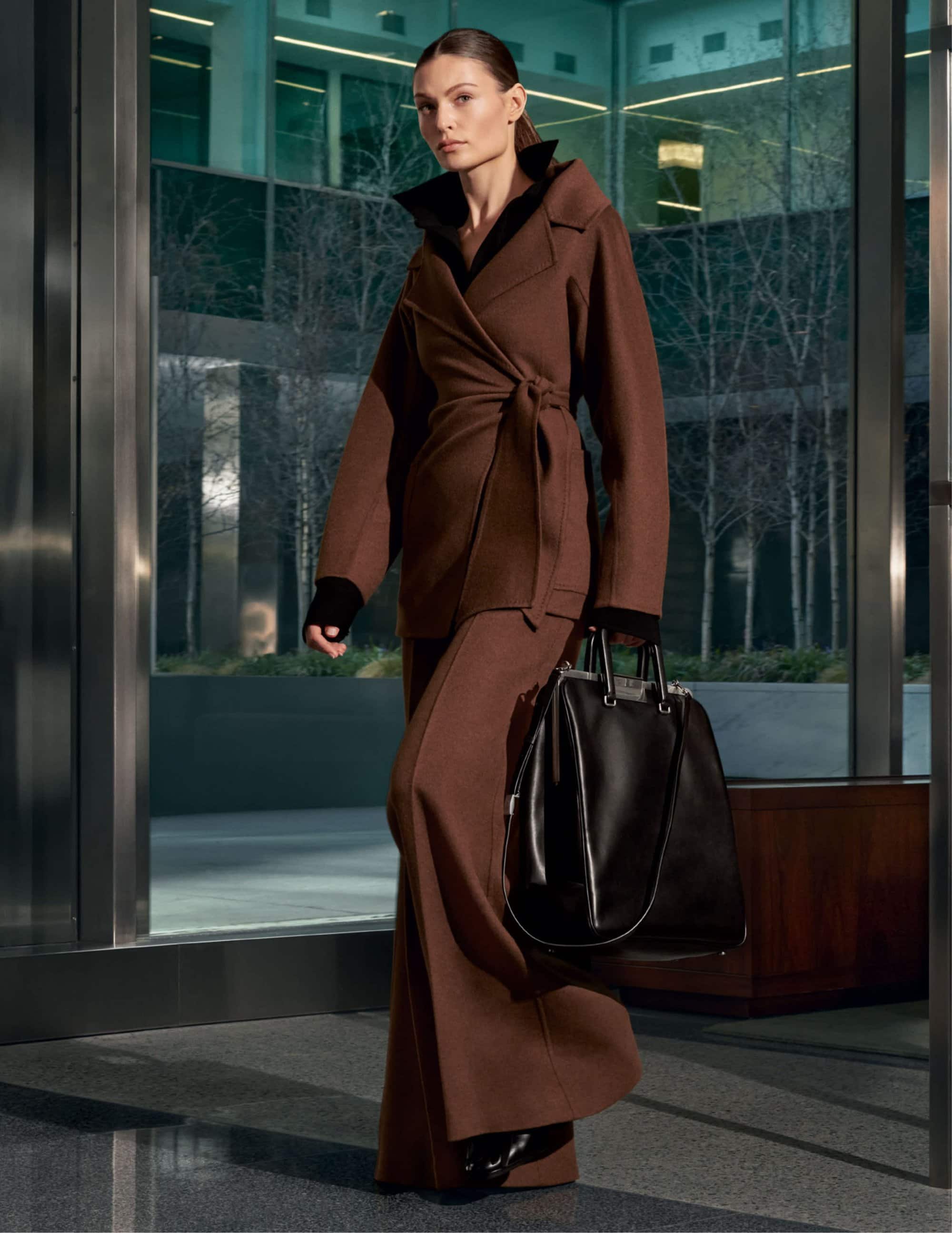 Model in brown belted coat and trousers holding a black handbag inside a modern building lobby with glass exterior
