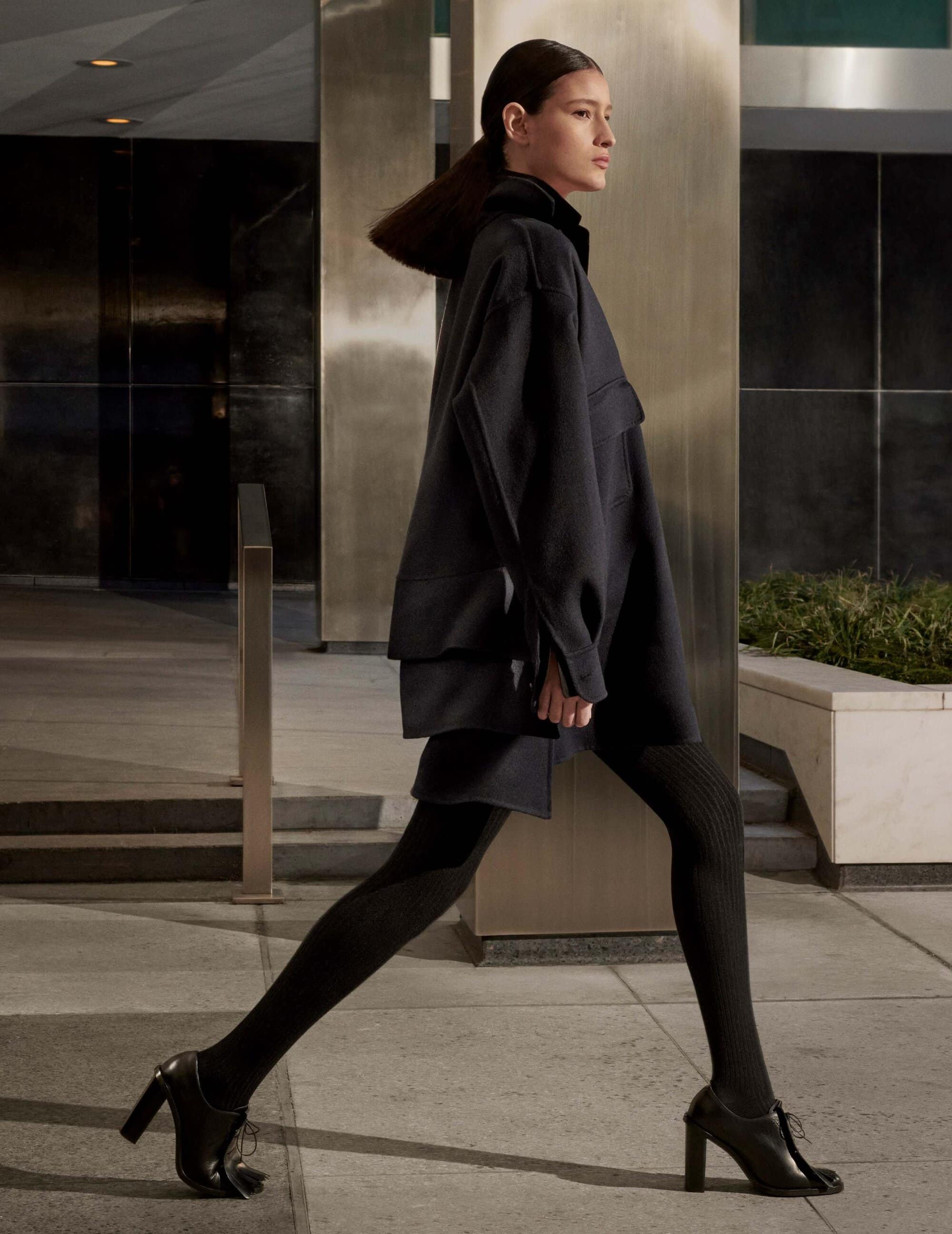 Model walking outdoors wearing a dark jacket, ribbed tights, and black high-heeled shoes