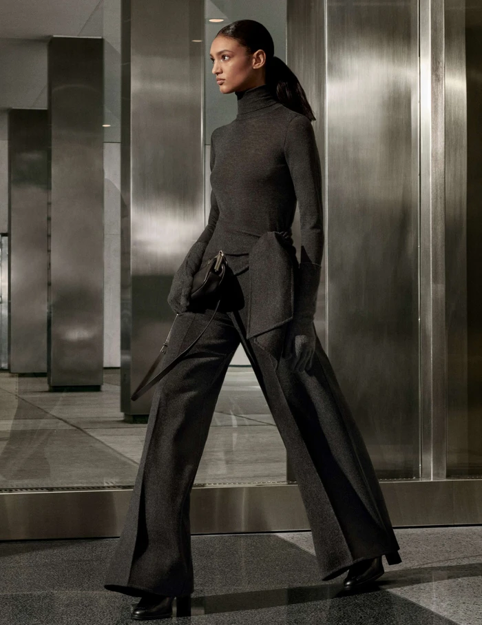 Model in charcoal grey turtleneck and wide trousers holding a handbag against a modern metallic architectural interior.