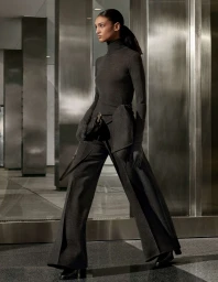 Model in charcoal grey turtleneck and wide trousers holding a handbag against a modern metallic architectural interior.