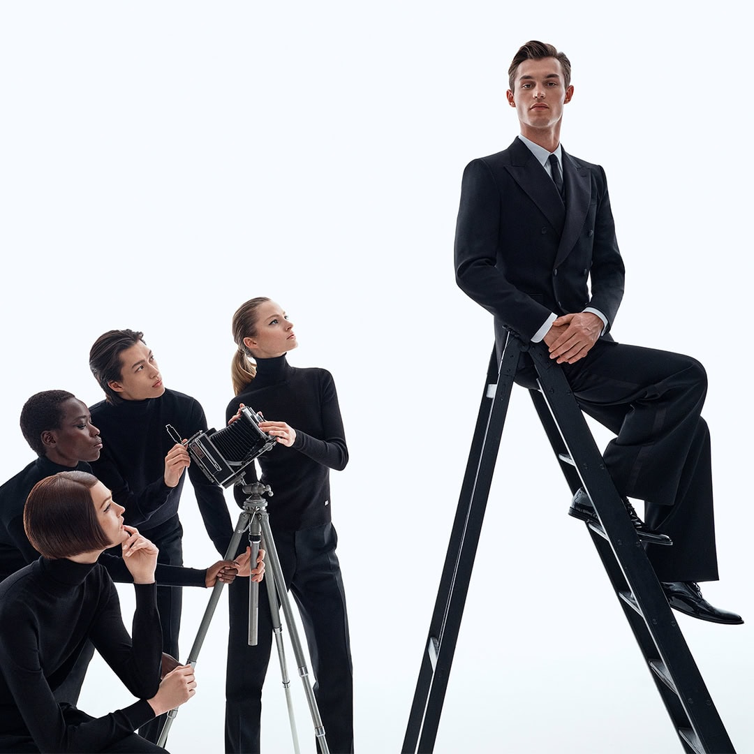 Man in black suit seated on a tall ladder with four people in black turtlenecks operating a vintage camera on a white background
