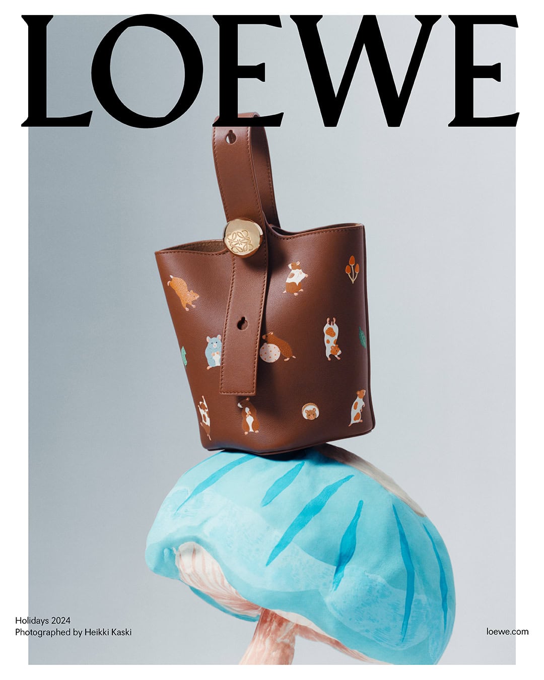 Brown leather bucket bag with animal illustrations resting on top of a light blue sculpted object against a pale grey background.