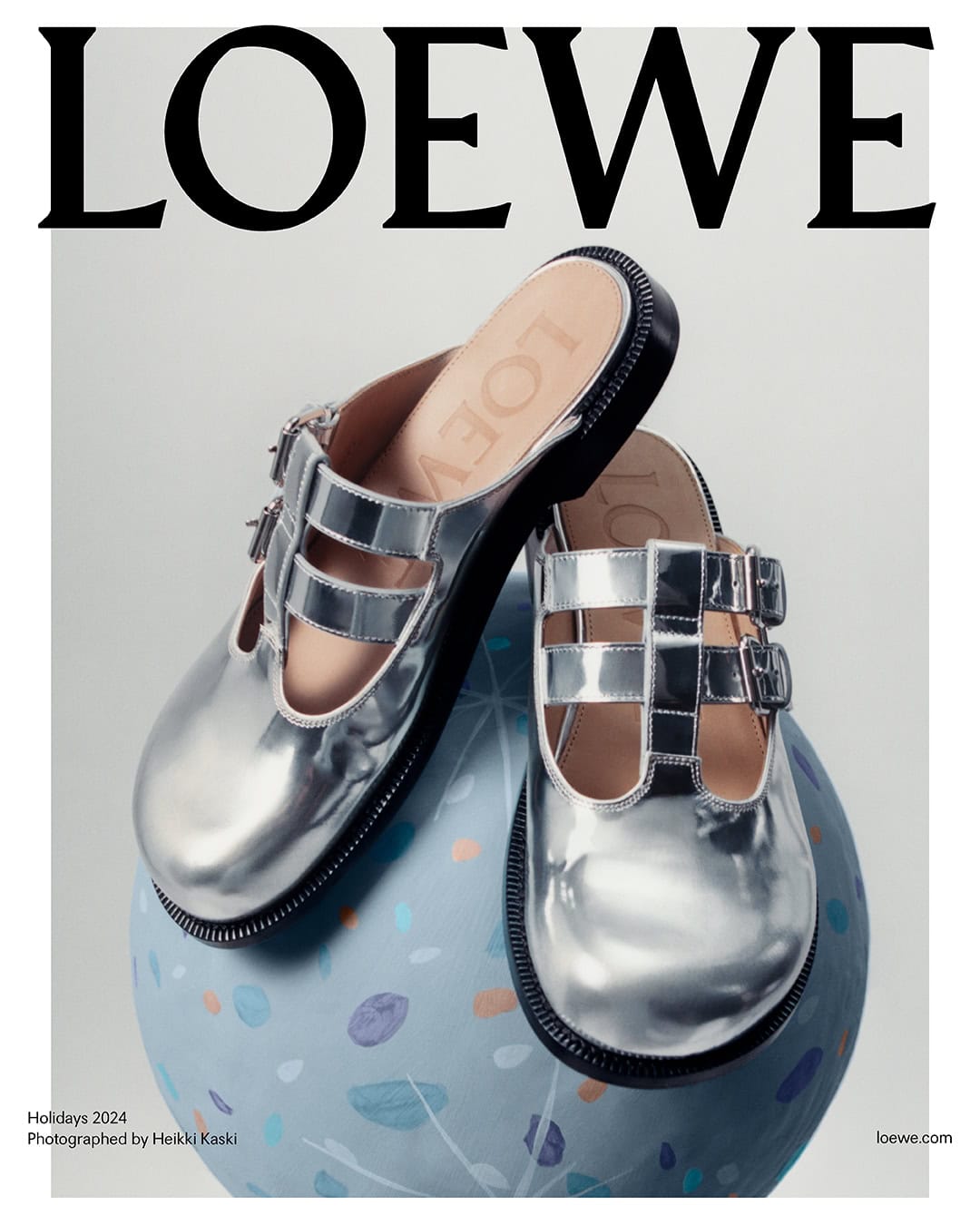 Pair of shiny silver buckled mules resting on top of a light blue decorative sphere against a neutral background