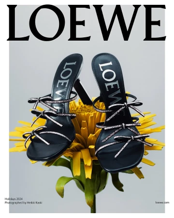 Black heeled sandals with crystal-embellished straps resting on a large yellow flower against a plain light grey background