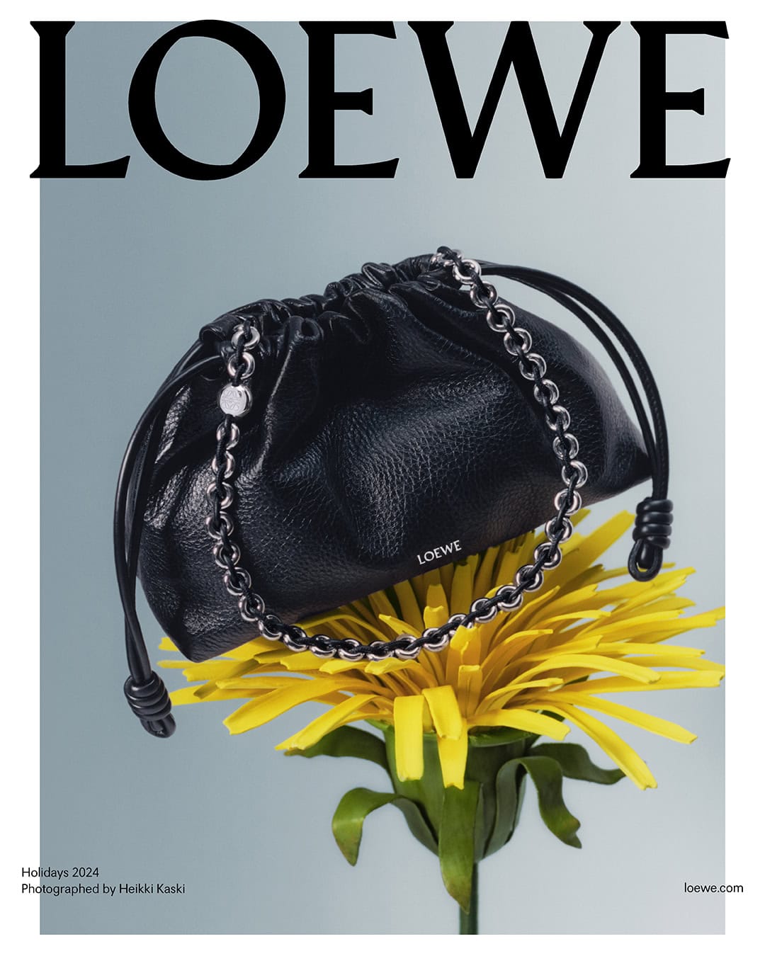 Black leather Loewe handbag resting on a bright yellow dandelion flower against a pale grey background
