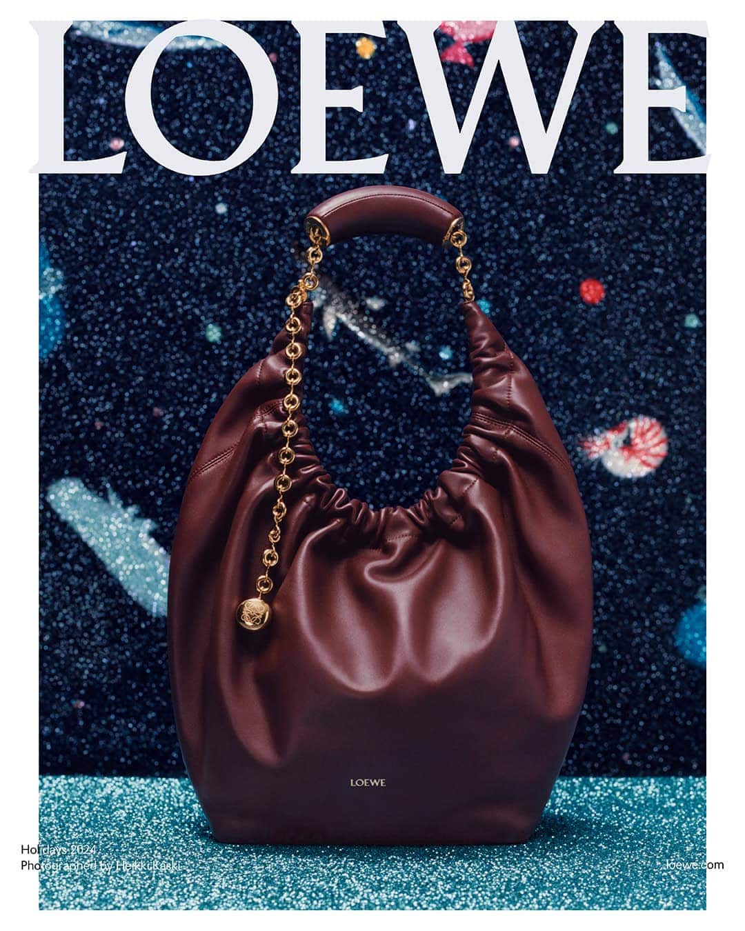 Burgundy leather hobo handbag with gold chain strap resting on a sparkly blue surface against a starry night background