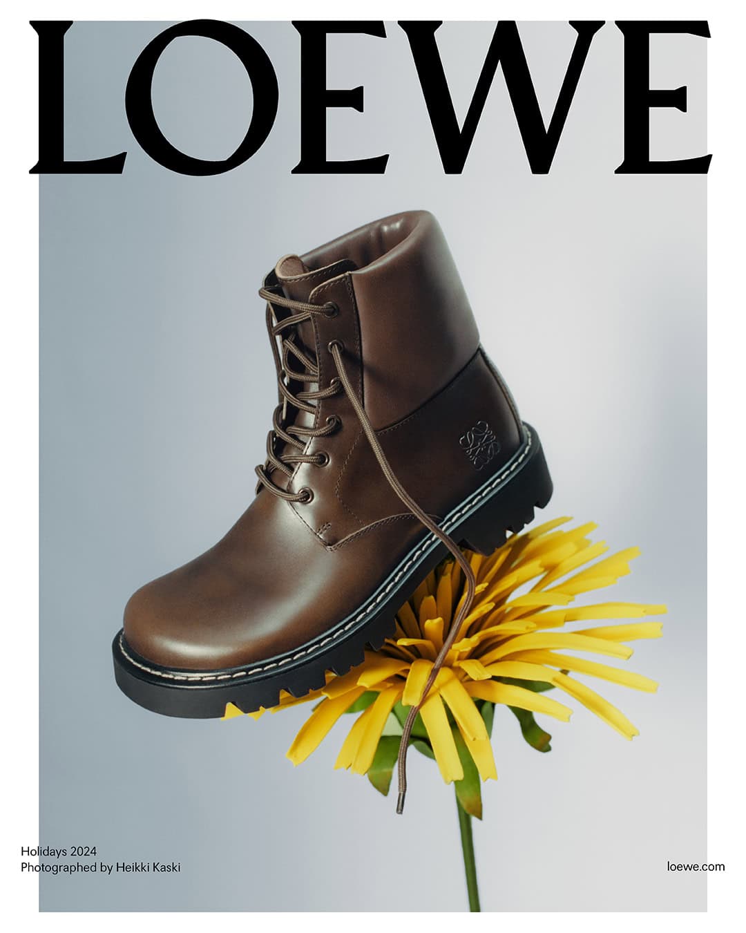 Brown leather boot resting on top of a bright yellow dandelion flower against a pale gradient background