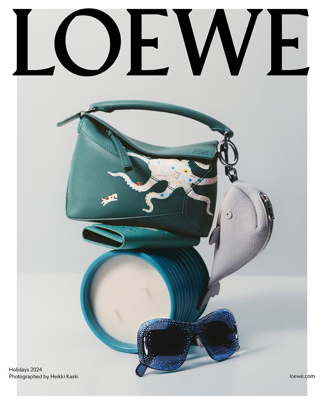 Loewe green leather handbag stacked on a candle and wallet with a whale charm and sunglasses against a white background