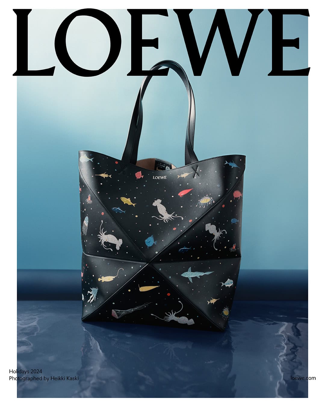 Black leather tote bag with colorful marine life illustrations set against a blue gradient studio backdrop with water reflection.