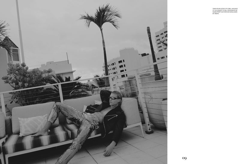 Black and white shot of a man reclining on a sofa on a terrace wearing sunglasses, a jacket, and patterned pants. Palm trees in the background.