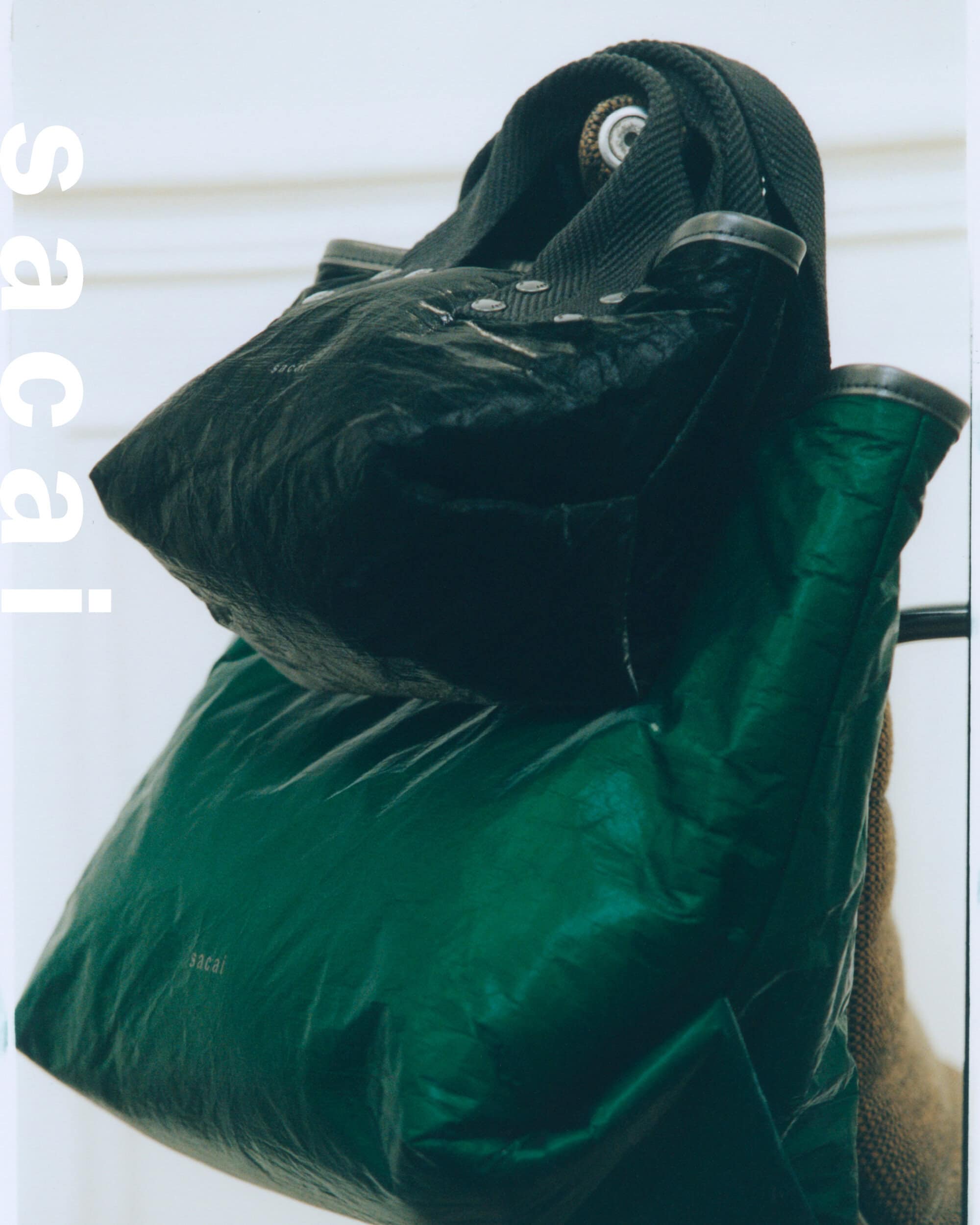 A black handbag stacked on top of a green nylon tote bag with white text against a neutral wall.