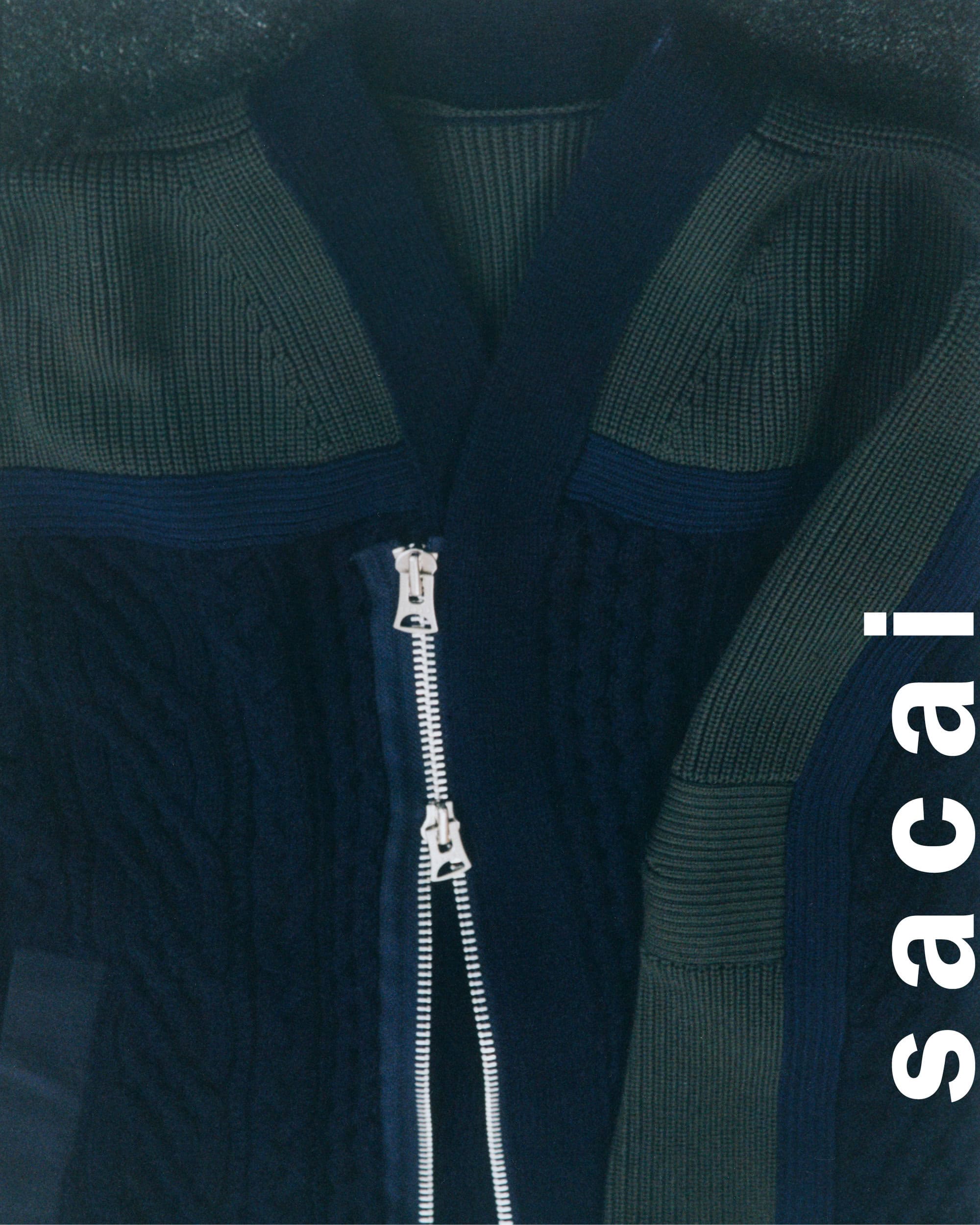 Close-up product shot of a navy blue and olive green knit cardigan with a prominent metal double zipper for Sacai.