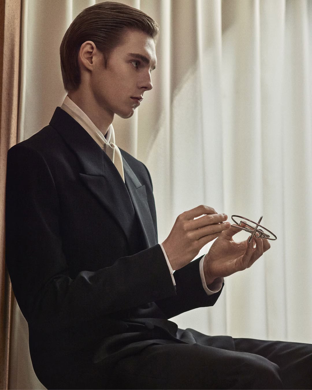 A man wearing a black suit jacket and white shirt holds a metallic object against a draped curtain background