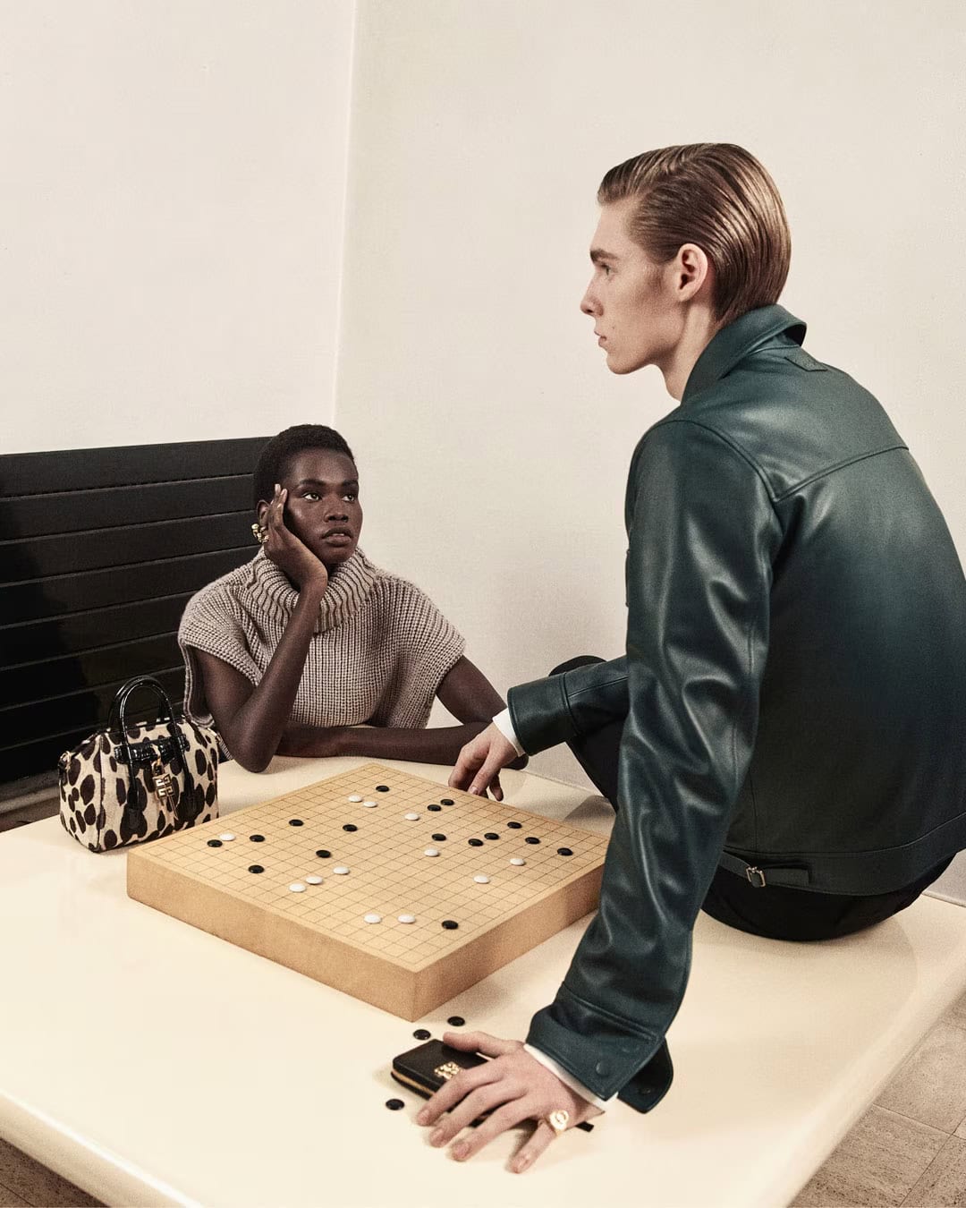 Two models playing a game on a wooden board with black and white pieces in a minimalist interior setting.