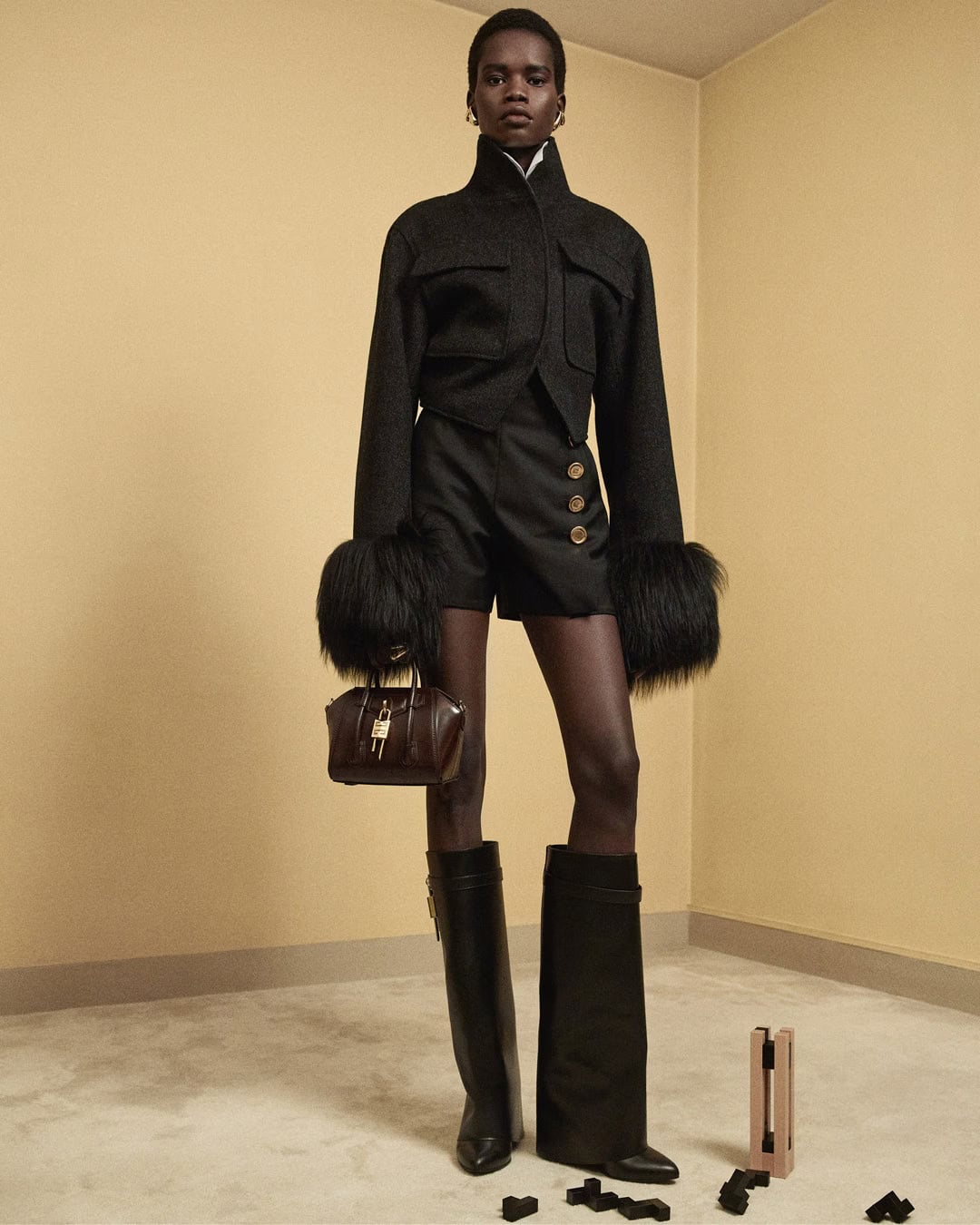 Model wearing a black jacket with fur cuffs, shorts, and tall black boots holding a small handbag against a beige wall.