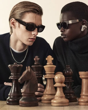 Two models wearing dark sunglasses and dark sweaters playing a game of chess for Givenchy Holiday 2024 campaign