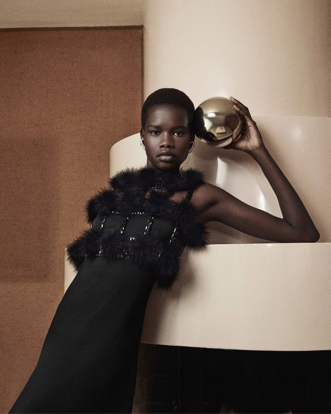 Model in black textured dress leaning against a curved architectural column holding a metallic sphere