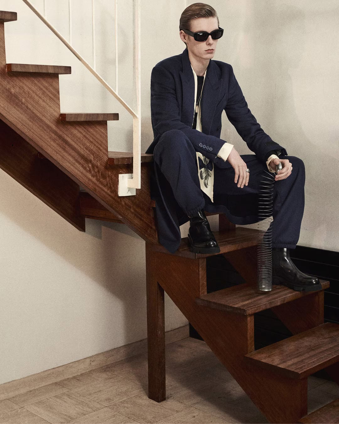 Man wearing dark sunglasses and a navy blue suit sitting on wooden stairs holding a metal spring toy