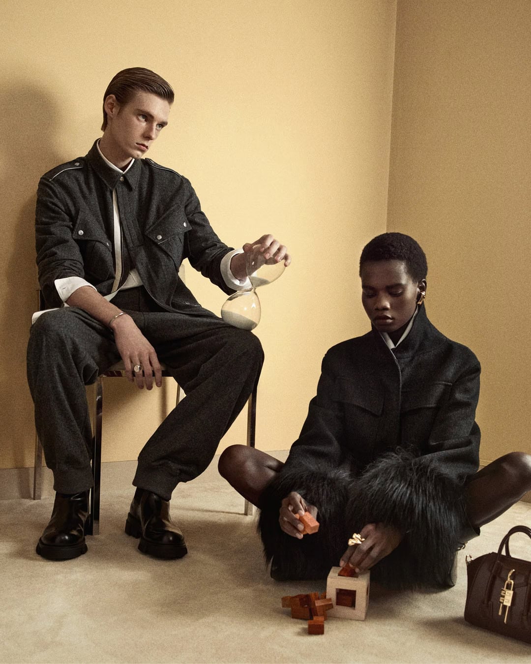 Two models in dark matching outfits pose inside a warm-toned room, with a man holding an hourglass and a woman playing with blocks