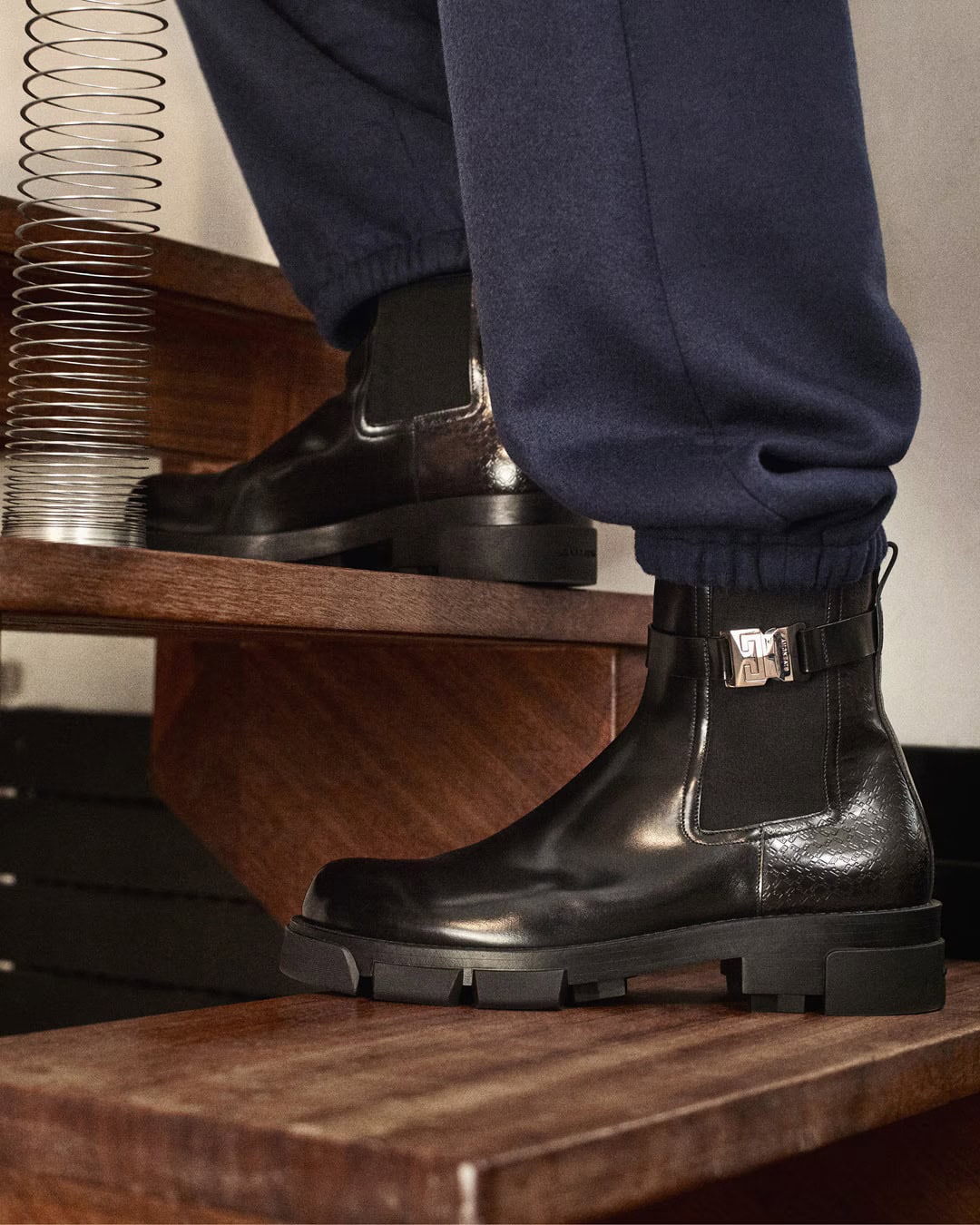 Close-up of black leather boots on dark trousers standing on wooden steps next to a metallic spring prop.
