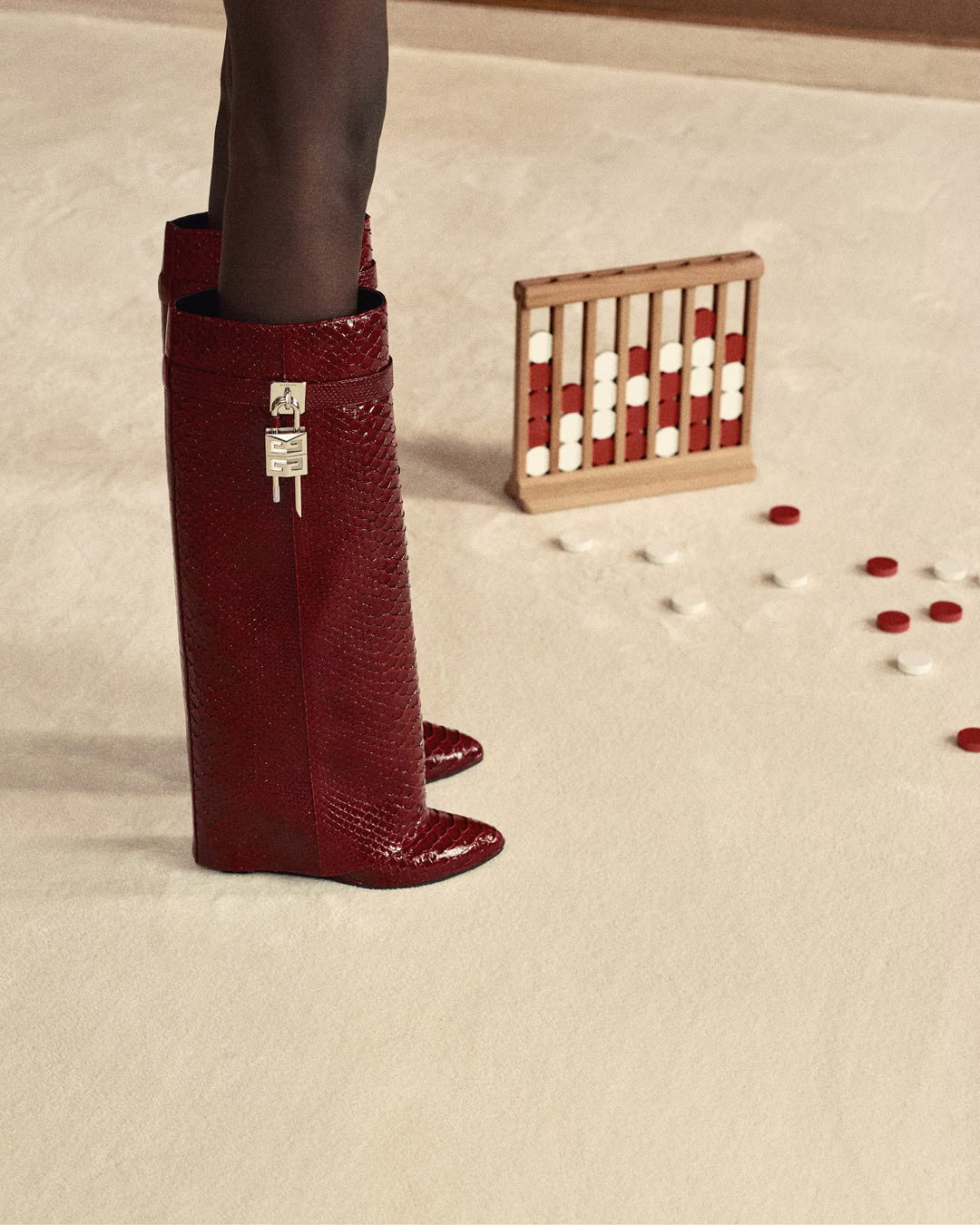 Close-up of tall red snakeskin boots with silver padlock hardware on a pale carpet near a wooden game set