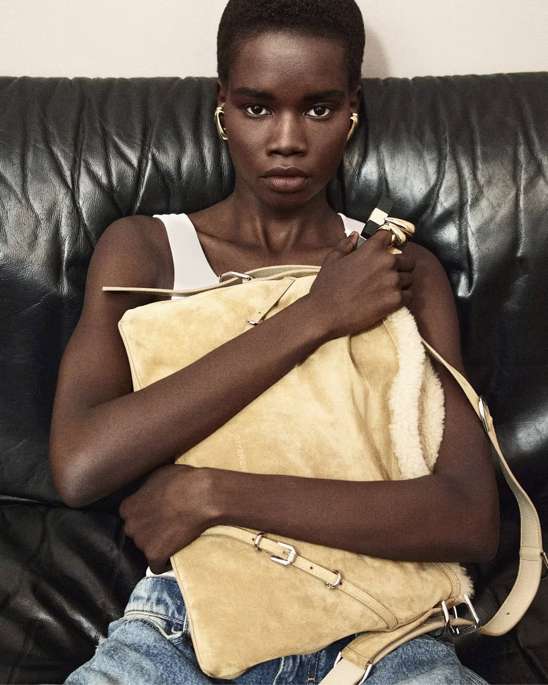 Model seated against a dark leather couch holding a large suede handbag for Givenchy.