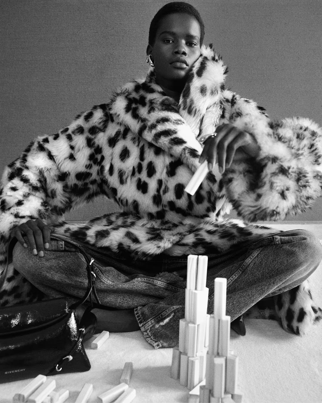 Model seated cross-legged wearing a spotted faux fur coat and denim jeans next to white block props and a black leather handbag