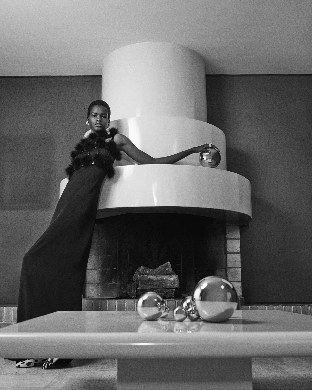Model wearing dark dress leaning against a large tiered fireplace structure in an interior setting.