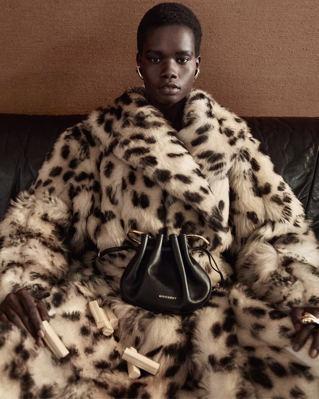 Model wearing a faux fur coat holding small objects with a black leather handbag resting in her lap