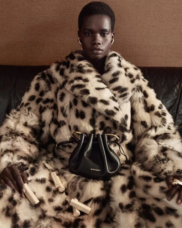 Model wearing a faux fur coat holding small objects with a black leather handbag resting in her lap