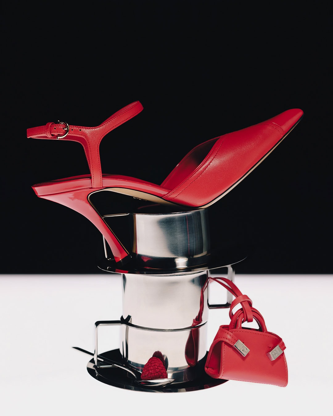 Red high heel shoe and miniature red handbag balanced on a stack of metal cups against a two-tone black and white background.